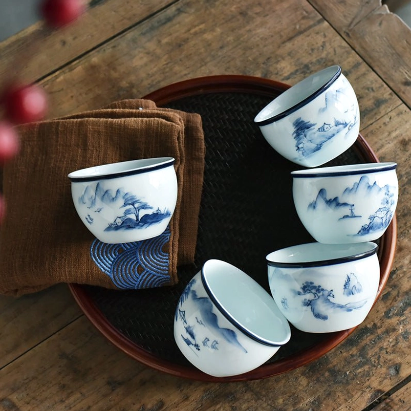 Multiple blue and white porcelain gongfu tea cups with mountain landscape paintings, top-down view with woven tray