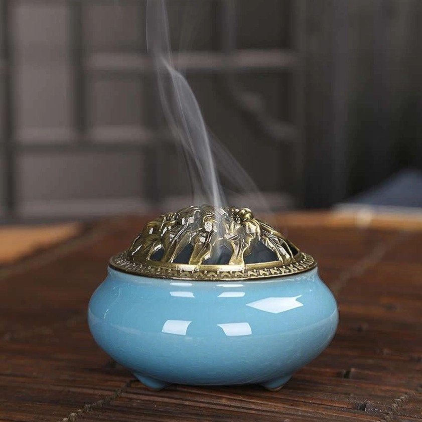 Hand-painted ceramic incense burner with rising incense smoke and decorative bronze floral lid on wooden table