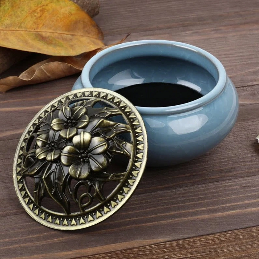 Open ceramic incense burner bowl with detachable bronze floral lid on wooden table with dry foliage