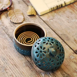 Handcrafted ceramic incense burner with separated floral carved teal lid and brown base, displayed on rustic wooden table with dried leaves and incense coil visible inside