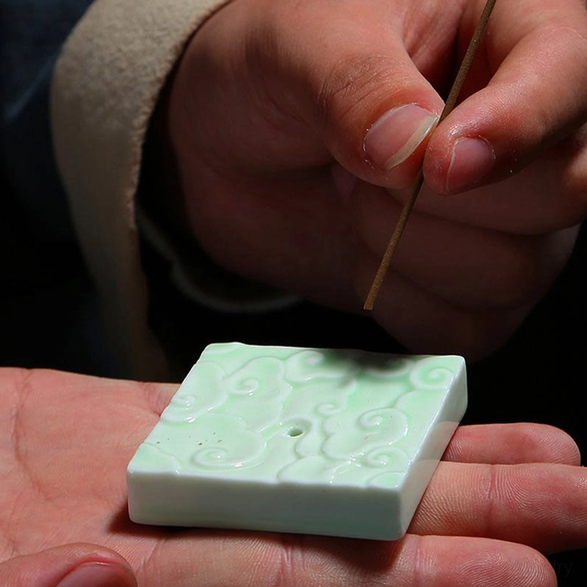 First-person perspective of inserting incense stick into ceramic cloud burner, displaying embossed cloud texture and single precision incense hole opening