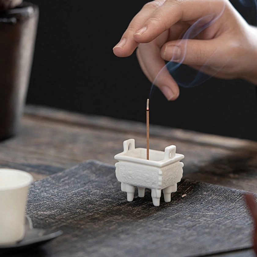 Small ceramic incense burner on tea table with dark placemat, hand placing incense stick showing meditation space setup