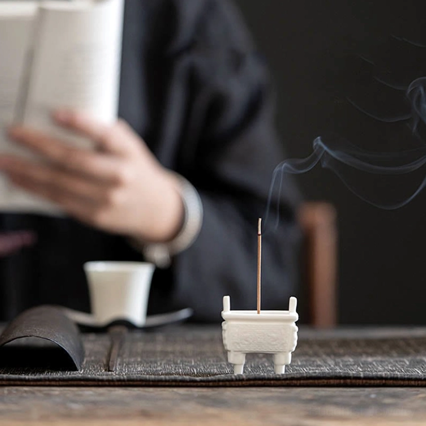 Buddhist ceramic incense burner on wooden table in reading space, burning incense with tea cup and peaceful indoor setting