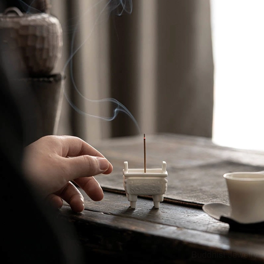 Mini ceramic incense burner on tea table with hand adjusting incense stick, curling aromatic smoke with tea set nearby