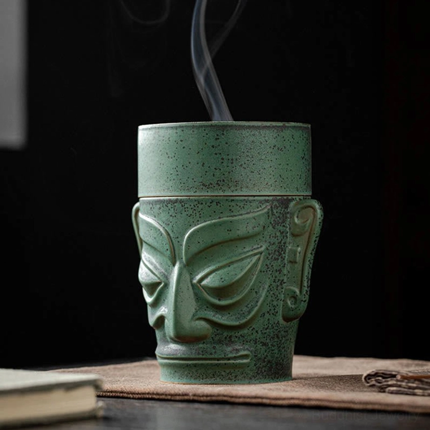 Left three-quarter view of ceramic mask incense burner with smoke emitting from top, carved facial mask details visible