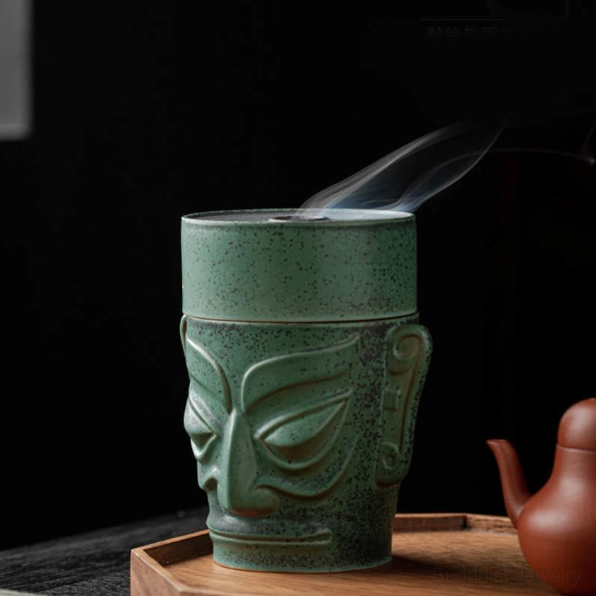 Low front three-quarter view of ceramic incense burner with curling smoke from top opening, muted green speckled glaze on wooden serving tray