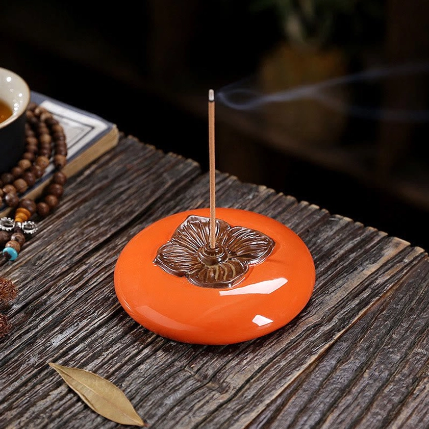 Handcrafted ceramic persimmon incense holder displayed on wooden table with prayer beads and dried leaf, showing bronze flower details