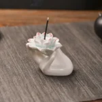 Handcrafted ceramic lotus hand incense burner with pink-tipped white lotus flower and light beige ceramic palm base, photographed from left three-quarter angle with burning incense stick
