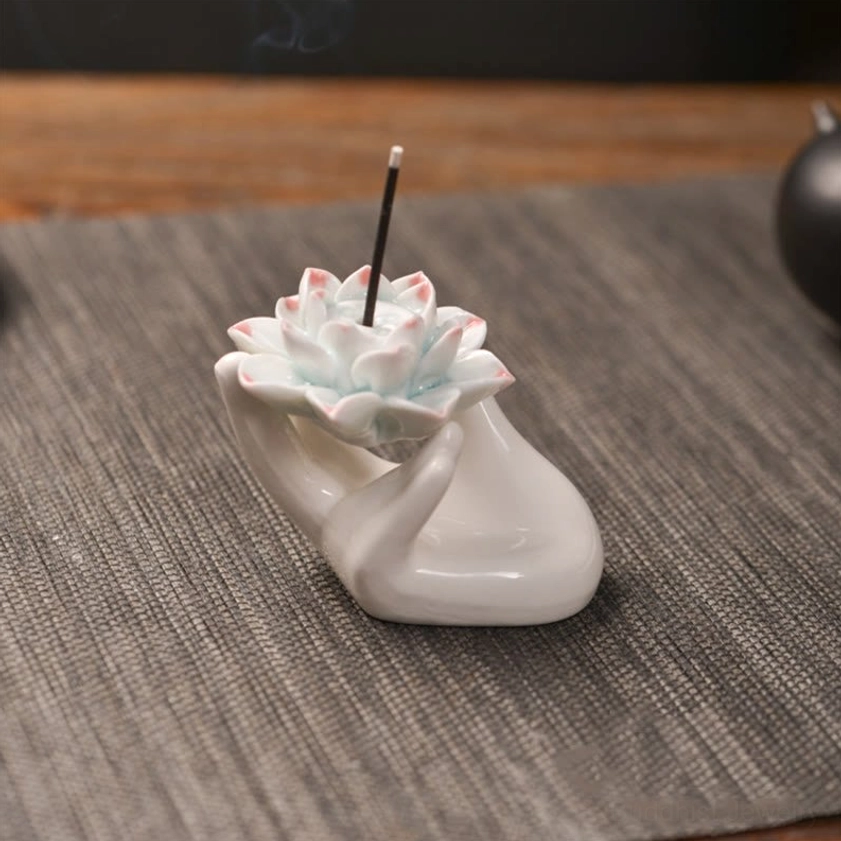 Handcrafted ceramic lotus hand incense burner with pink-tipped white lotus flower and light beige ceramic palm base, photographed from left three-quarter angle with burning incense stick