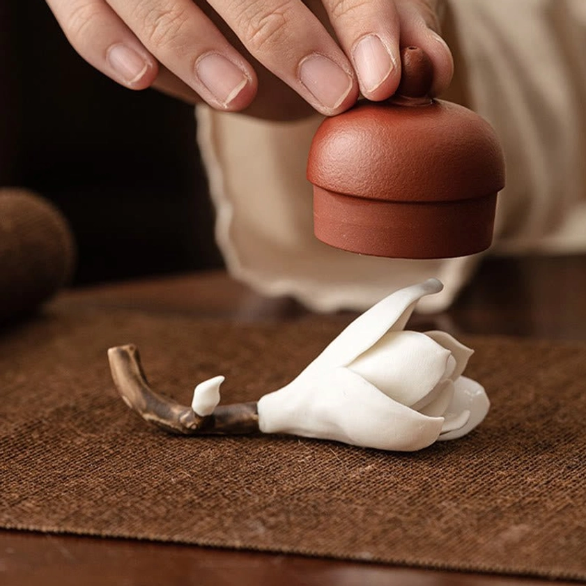 Handcrafted ceramic magnolia incense holder with lit incense on woven brown placemat in warm indoor setting near teapot