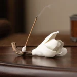 White ceramic magnolia incense holder with burning incense stick on dark wooden surface, showing detailed handcrafted flower design