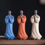 Three handcrafted ceramic meditation monk statues in blue, white, and terracotta colors with prayer bead necklaces displayed on wooden table
