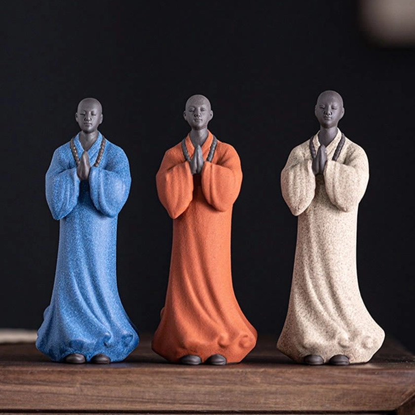 Three handcrafted ceramic meditation monk statues in blue, white, and terracotta colors with prayer bead necklaces displayed on wooden table