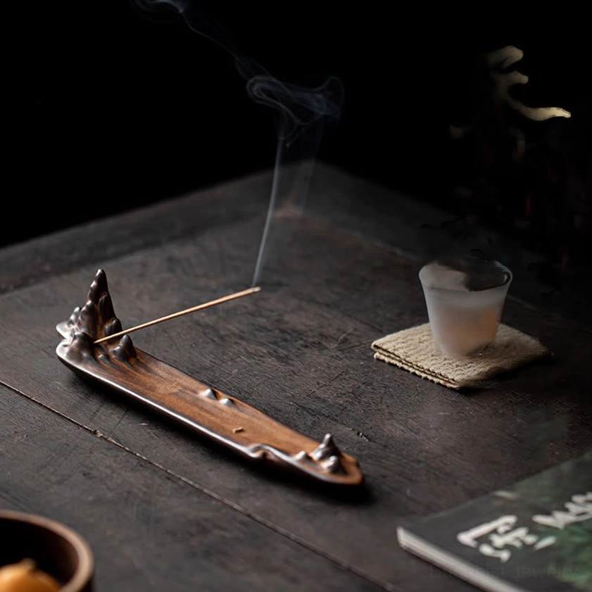 Low side angle of ceramic mountain incense stick burner on dark wooden table with tea accessories, showing curling incense smoke