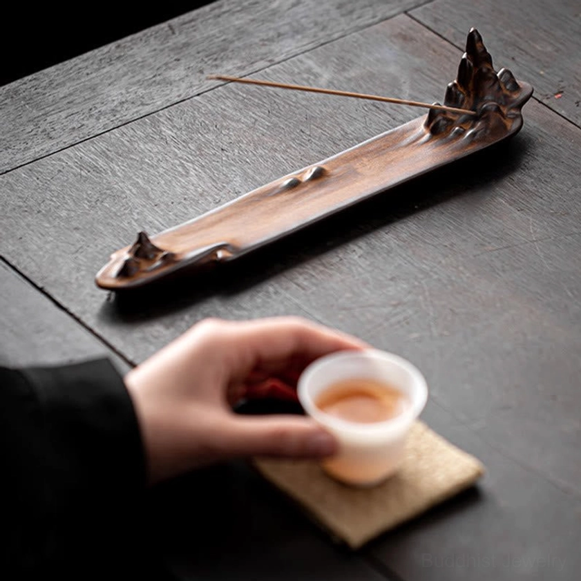 Close-up side view of ceramic mountain incense holder on dark wood tea table with tea cup and coaster nearby, showing incense placement