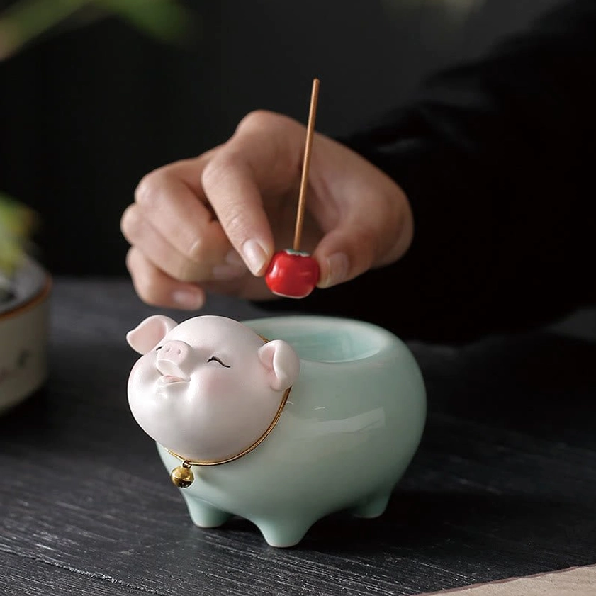 Close-up front view of ceramic pig incense burner on wooden table, hand placing red cherry decoration on smiling pig face