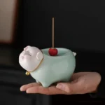 Handcrafted ceramic pig incense burner with celadon glaze held in palm, showing detailed pig face with gold bell collar and red cherry decoration