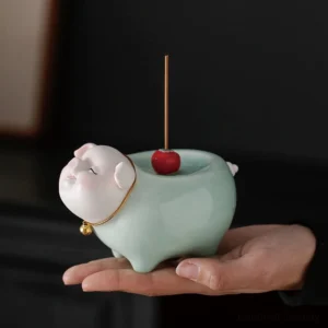Handcrafted ceramic pig incense burner with celadon glaze held in palm, showing detailed pig face with gold bell collar and red cherry decoration