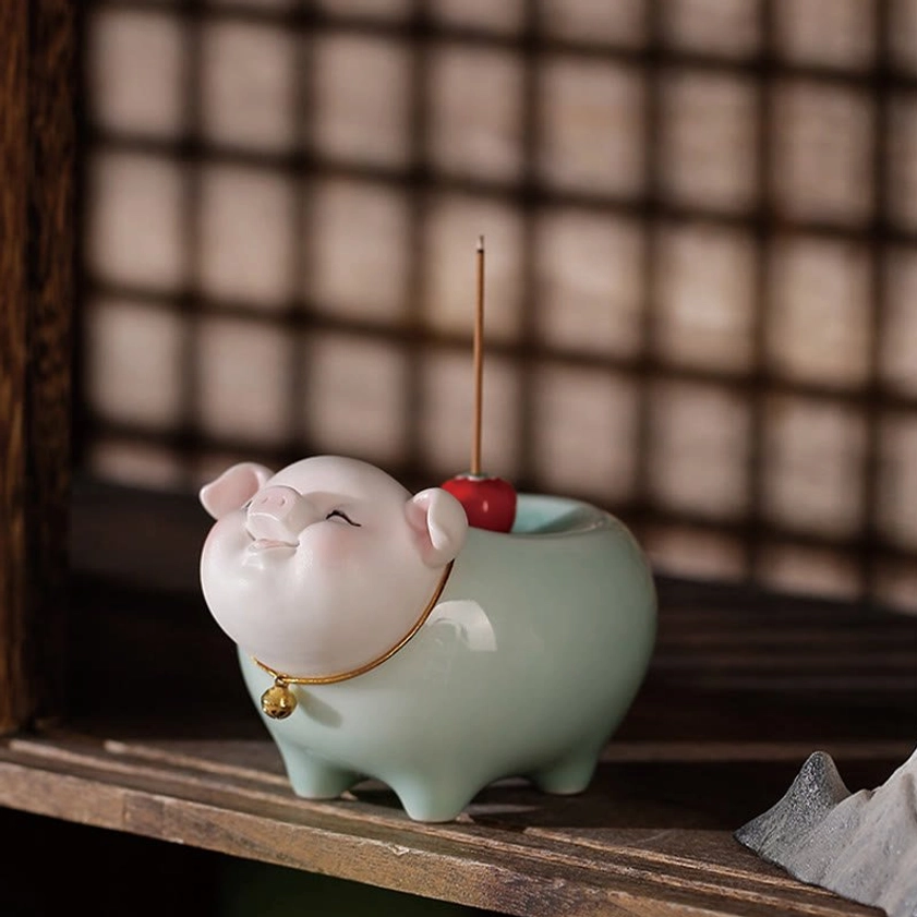 Ceramic pig incense burner displayed on wooden shelf with grid window background, gold bell collar clearly visible