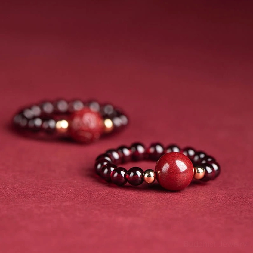 Two cinnabar accent beaded Buddhist rings with gold tone spacers and distinct hand-carved focal charm designs arranged on wine-red background, showcasing luxury details