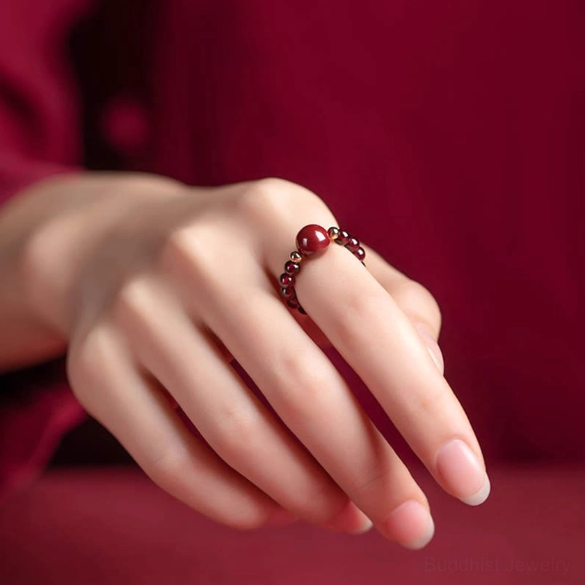 Hand wearing adjustable cinnabar beaded Buddhist ring with small red accent bead on middle finger, wine-red clothing backdrop showing natural hand positioning