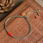 Handmade cinnabar double-layer braided bracelet with blue-yellow woven cord and red cinnabar charm, displayed on wooden table with dried flowers