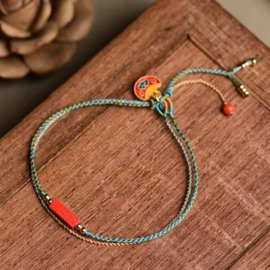 Handmade cinnabar double-layer braided bracelet with blue-yellow woven cord and red cinnabar charm, displayed on wooden table with dried flowers