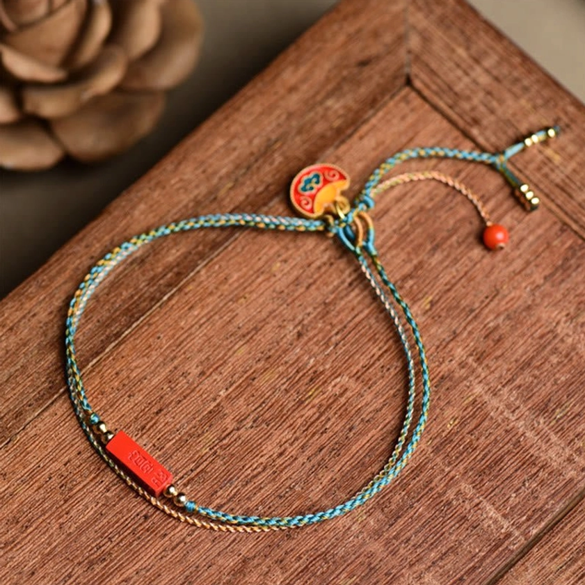 Handmade cinnabar double-layer braided bracelet with blue-yellow woven cord and red cinnabar charm, displayed on wooden table with dried flowers