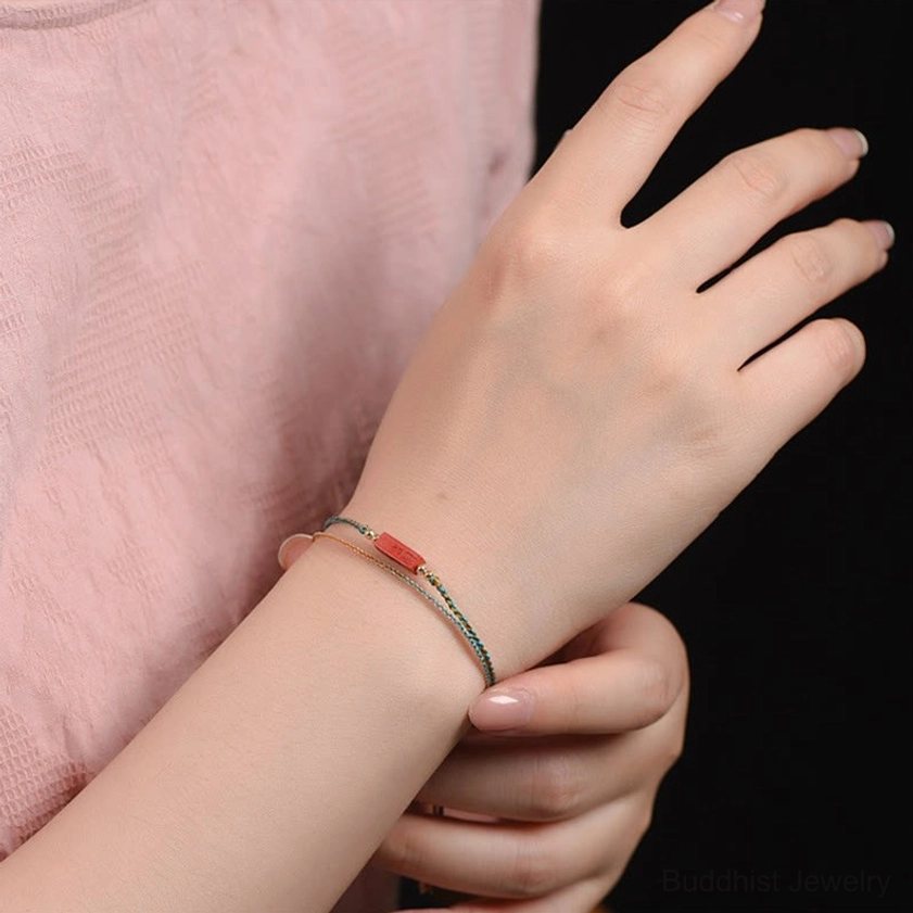 Handmade cinnabar braided bracelet worn on wrist, showcasing thin braided cord with delicate red cinnabar tag and metal accents