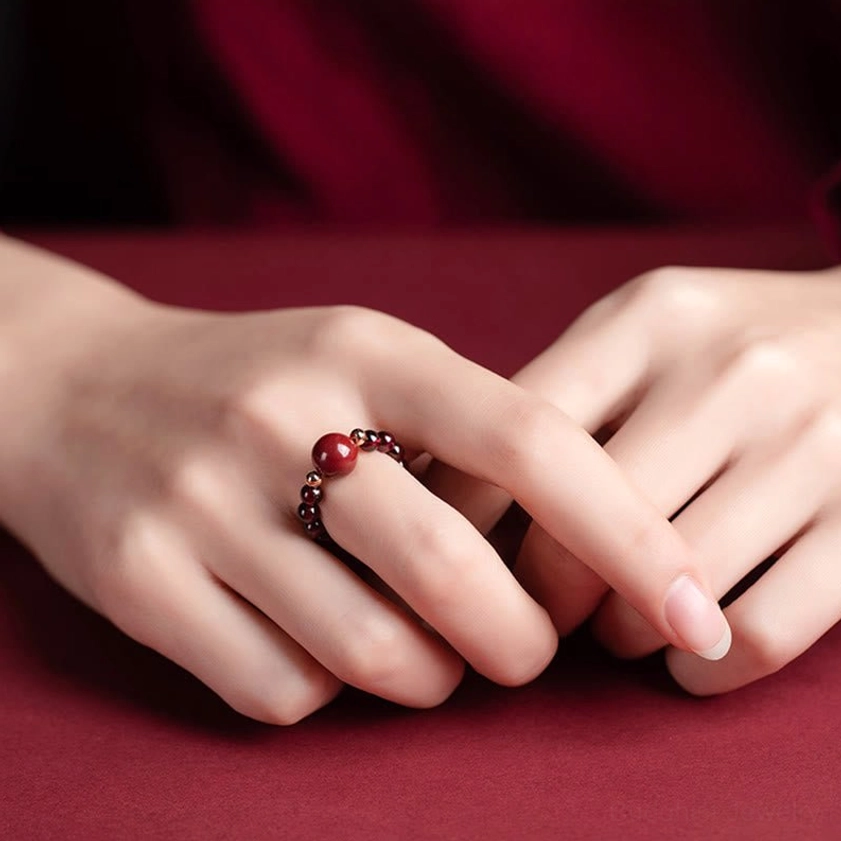Close-up hand wearing delicate cinnabar beaded Buddhist ring on middle finger with wine-red clothing backdrop, showing hand-carved details and adjustable fit