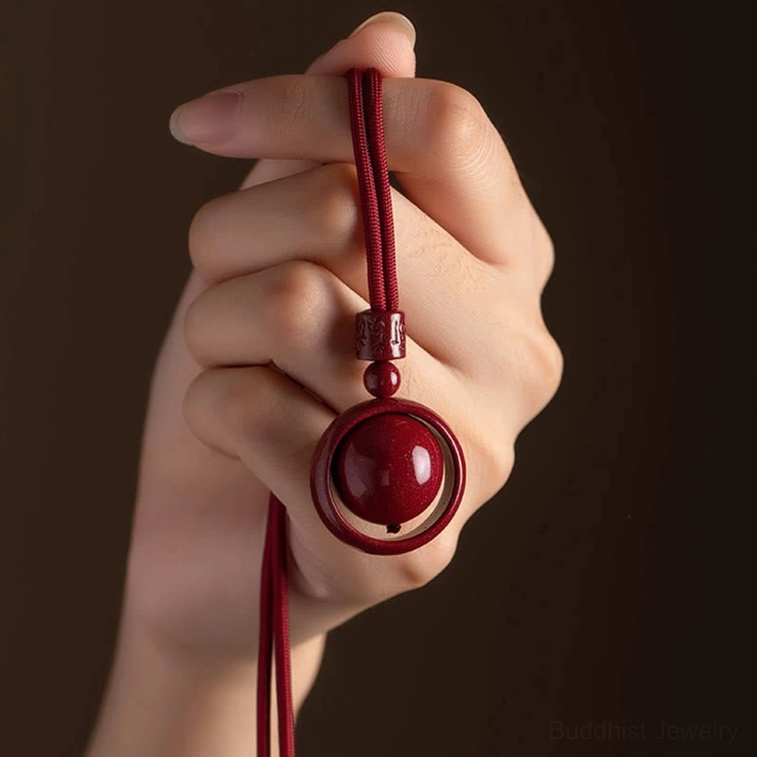 Handmade cinnabar round bead pendant with hollow ring held against dark background, red woven cord