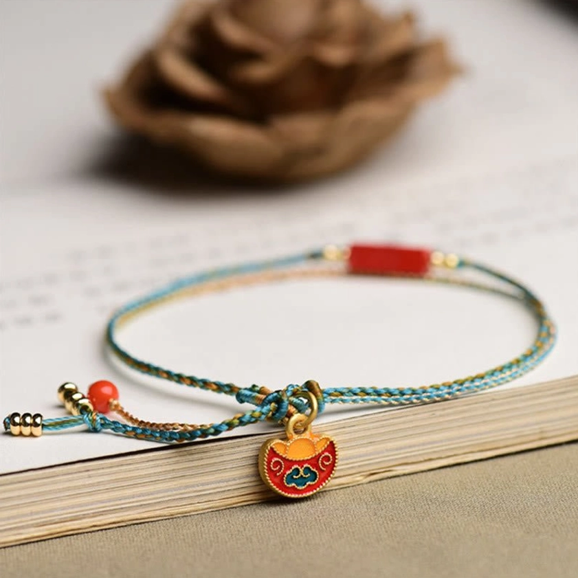 Woven cinnabar bracelet with lock-shaped enamel pendant and red tassel on open book desktop, side-top view