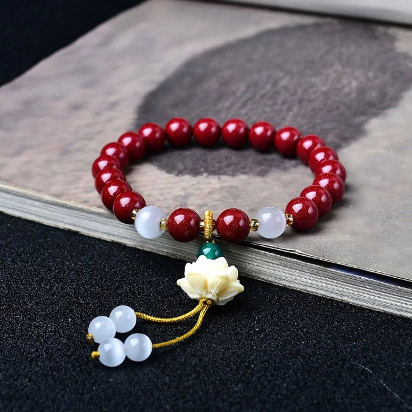 Handcrafted cinnabar lotus blessing bracelet featuring deep red cinnabar beads, white ivory fruit spacers, carved lotus charm pendant, and golden tassel - displayed on vintage book