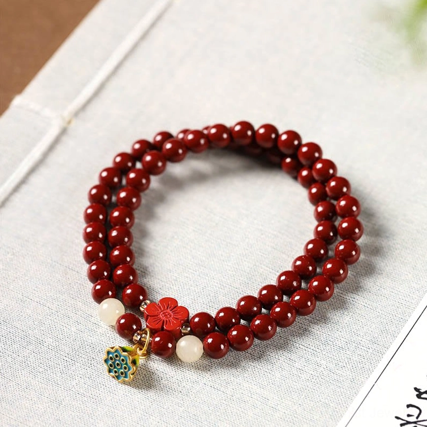 Cinnabar lotus double-wrap blessing bracelet with red beads and enamel lotus charm on linen notebook flat lay