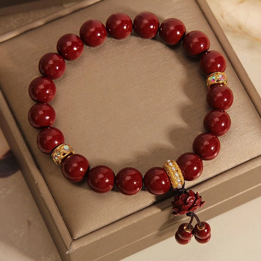 Top-down flat lay of cinnabar lotus blessing bracelet on brown jewelry tray, showing maroon beads, gold crystal spacers, and red lotus charm with bead tassel