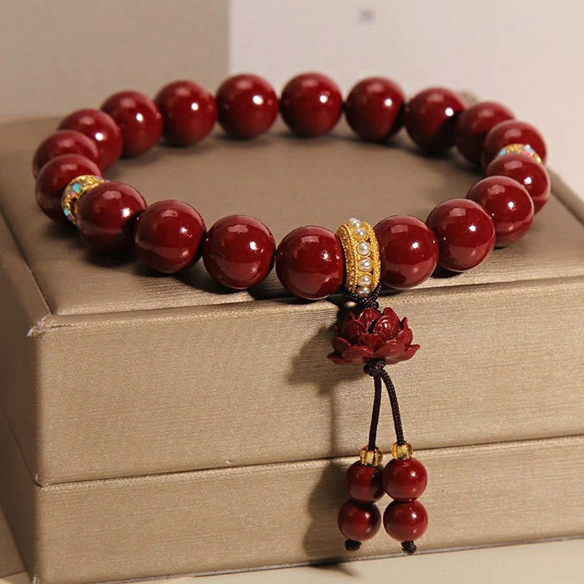 Four-quarter view of cinnabar lotus blessing bracelet stacked on brown jewelry boxes, showing dark red polished beads, gold crystal accents, and knotted lotus tassel