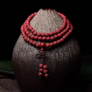 Carved cinnabar 108 mala beads bracelet with engraved Om Mani Padme Hum pattern, displayed on dark ceramic vase