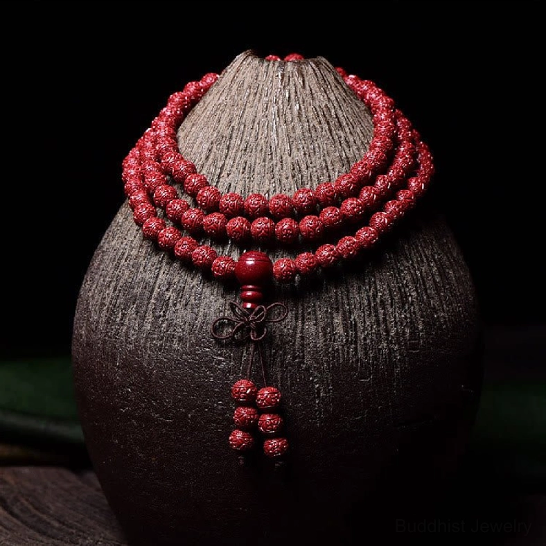 Carved cinnabar 108 mala beads bracelet with engraved Om Mani Padme Hum pattern, displayed on dark ceramic vase