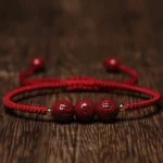Handmade cinnabar Om Mani Padme Hum engraved beads braided bracelet with dark red cord, flat lay on wooden surface showing carved mantra details and copper spacer beads