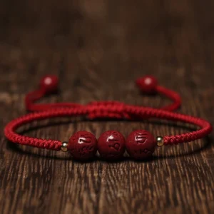 Handmade cinnabar Om Mani Padme Hum engraved beads braided bracelet with dark red cord, flat lay on wooden surface showing carved mantra details and copper spacer beads