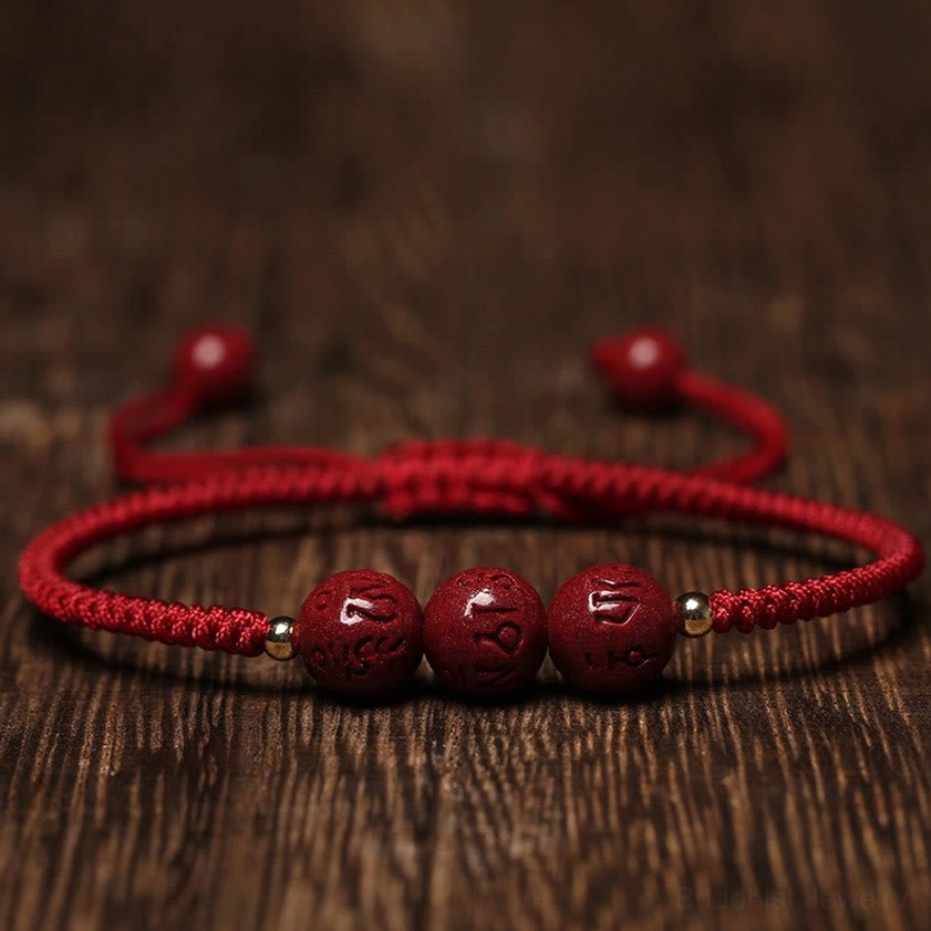 Handmade cinnabar Om Mani Padme Hum engraved beads braided bracelet with dark red cord, flat lay on wooden surface showing carved mantra details and copper spacer beads