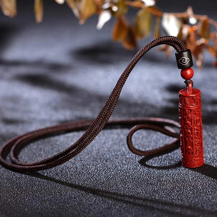 Cinnabar mantra pendant necklace laid flat on dark textured tabletop, showing cylindrical carved pendant with brown hanging cord
