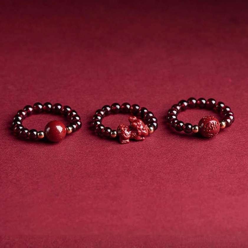 Three cinnabar Buddhist meditation rings with PiXiu charm, carved beads, and large focal cinnabar beads arranged flat on wine-red background, displaying different spiritual charm variations