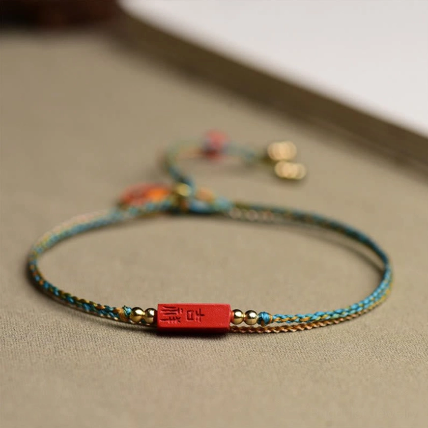 Rectangular cinnabar Ji Xiang plaque bracelet with blue-yellow braided cord and gold spacer beads on neutral fabric