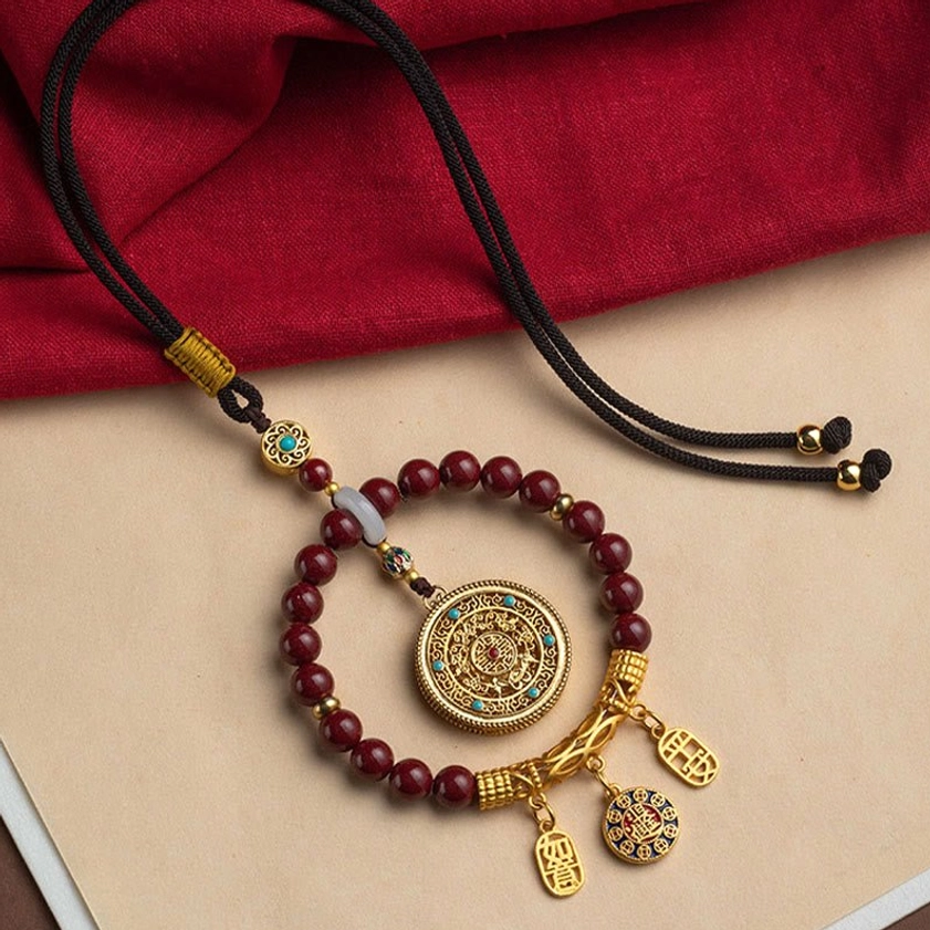 Handmade cinnabar blessing ornament displayed on red fabric with adjustable black braided cord, showing intricate gold filigree detail