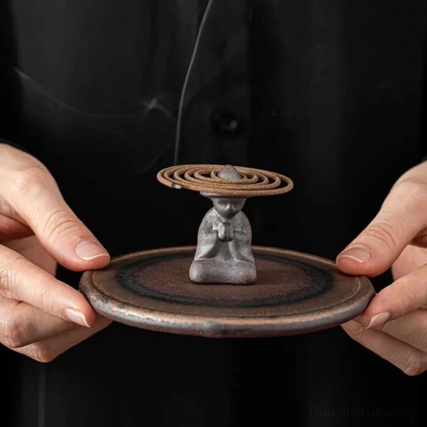 Glazed ceramic coil incense burner with monk figurine held in hands against dark background, showcasing artisan craftsmanship