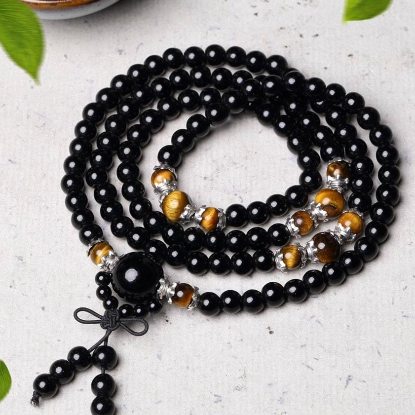Coiled 108 bead black obsidian Buddhist mala with central large bead, tiger eye stones, and tassel pendant on dark table with green leaf