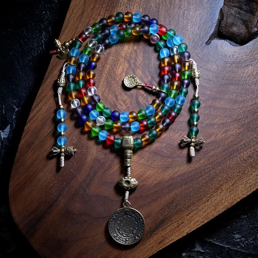 Complete 108-bead Buddhist mala set with mixed color Liuli glass beads, multiple metal charms including round coin pendant, flat lay on wooden board
