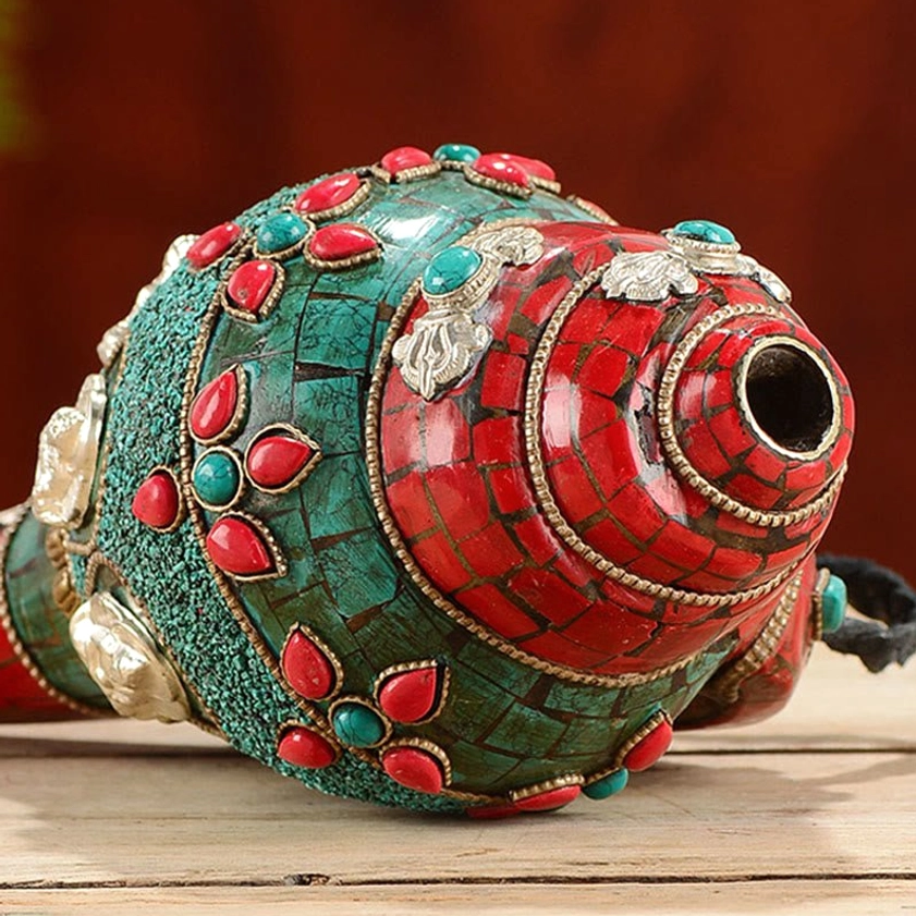 Close-up side view of handcrafted Buddhist conch shell with copper spout, turquoise and gemstone mosaic inlays against dark red background