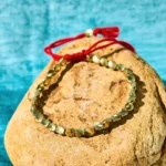 Handcrafted copper bead bracelet with red tassel closure displayed on stone background, matte gold finish for Buddhist protection and prosperity